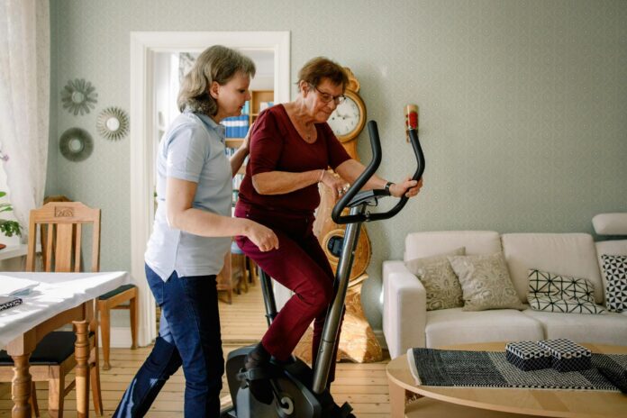 Mature female physical therapist assisting elderly woman on exercise bike at nursing home